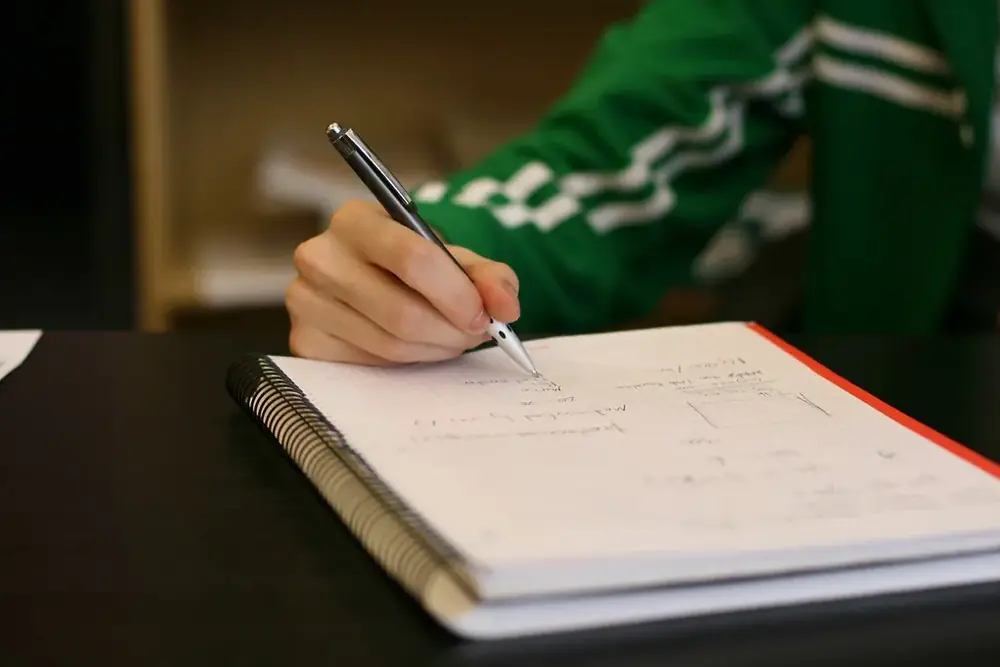 A hand with a pen writing calculations on a spiral notebook page
