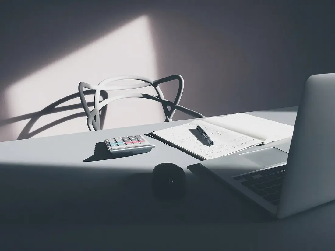 A pale desk with a notebook, a pen, a calculator and a laptop under a shaft of window light