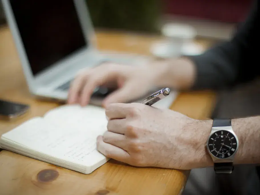 A person writing in a small notebook beside an open laptop on a light wooden desk