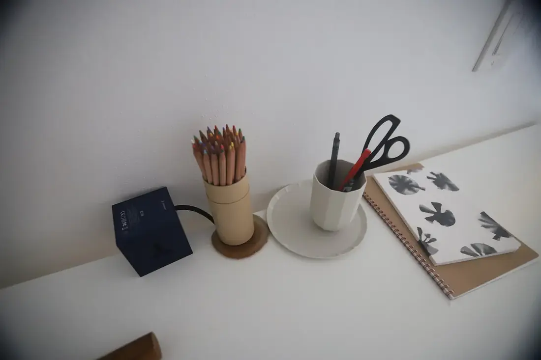A white desk with a pot of coloured pencils, a cup of pens, scissors and a spiral notebook