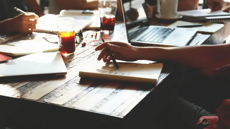 Four people at a wooden table with notebooks, glasses of iced tea and an open laptop; one hand writes in a notebook